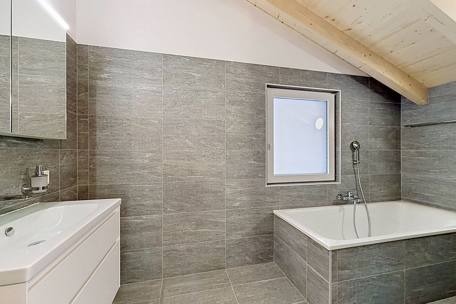 Salle de bains neuve : baignoire, carrelage gris pierre, meuble vasque blanc, fenêtre, charpente apparente.