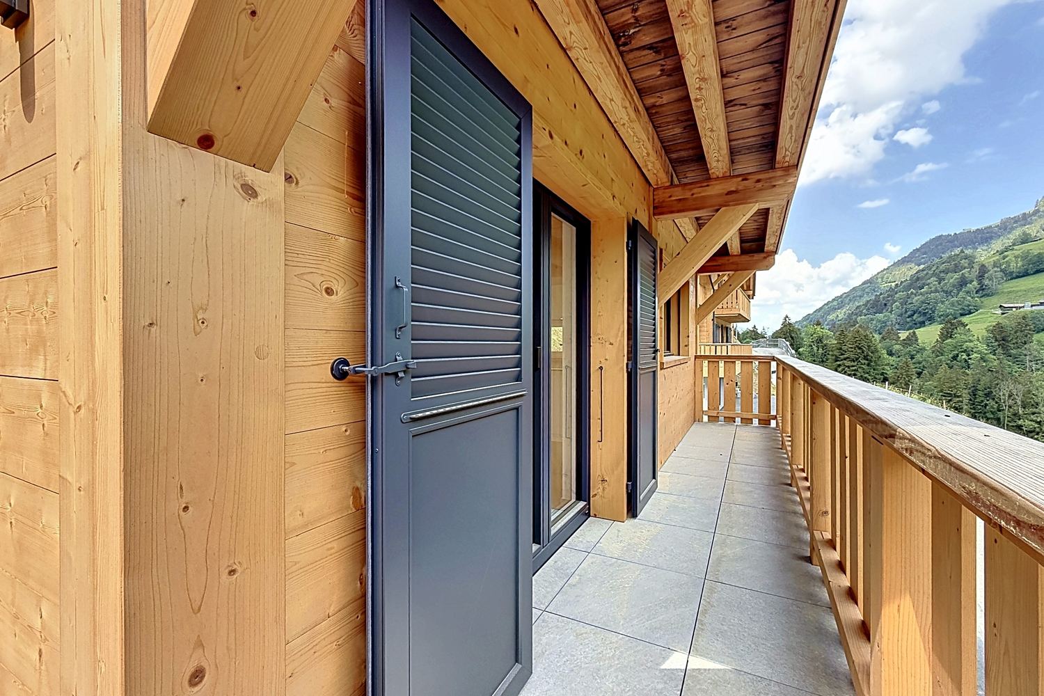 Long balcon de bois à l'étage des chambres, volets bleu-gris, avant-toit et vue sur les montagnes.