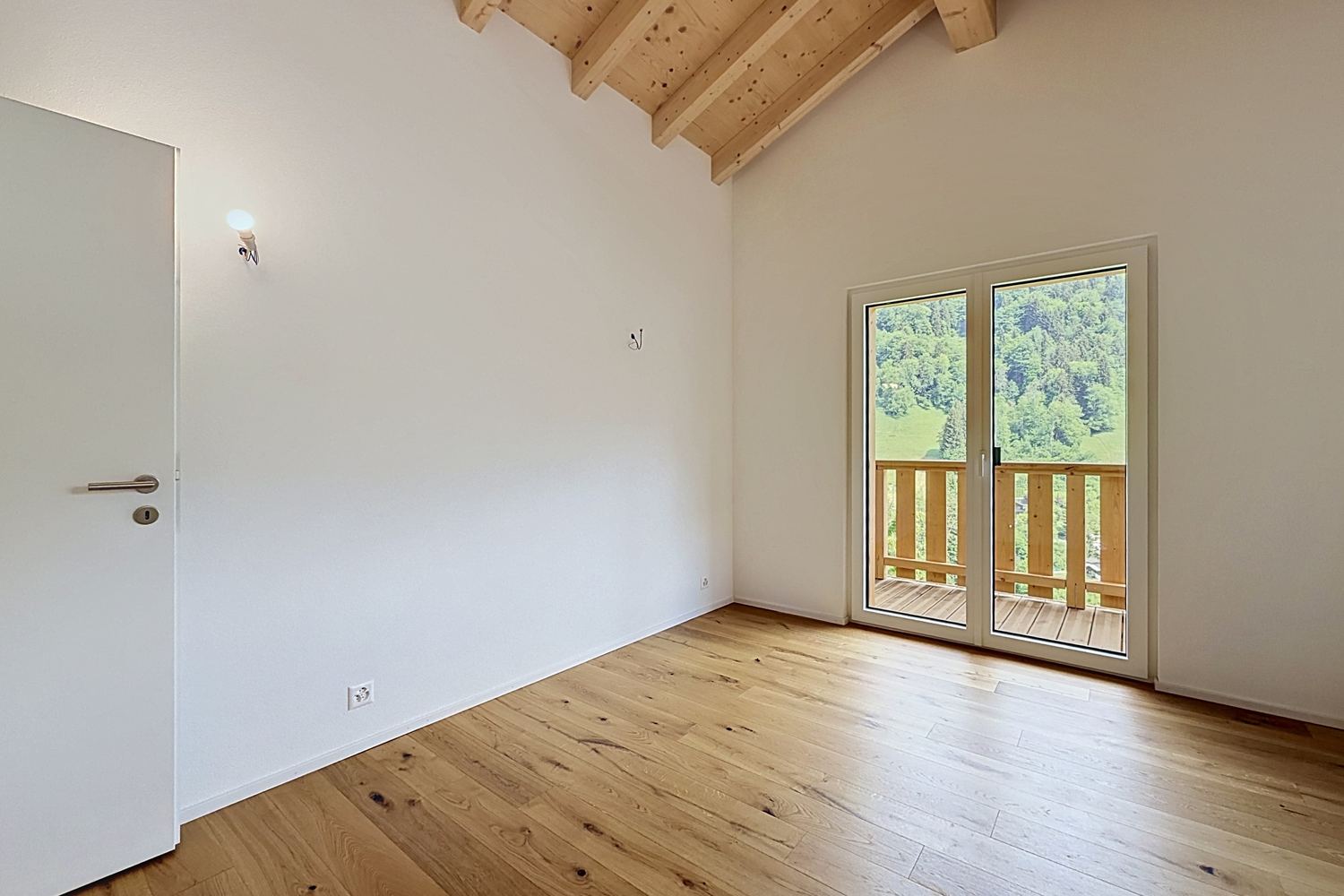 Chambre vide sous les combles, parquet de chêne, porte-fenêtre ouvrant sur le balcon et la forêt.