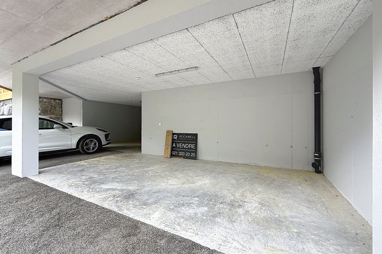 Couvert à voiture en béton sous le chalet, voiture garée, tableau électrique au mur, ouverture sur l'accès.