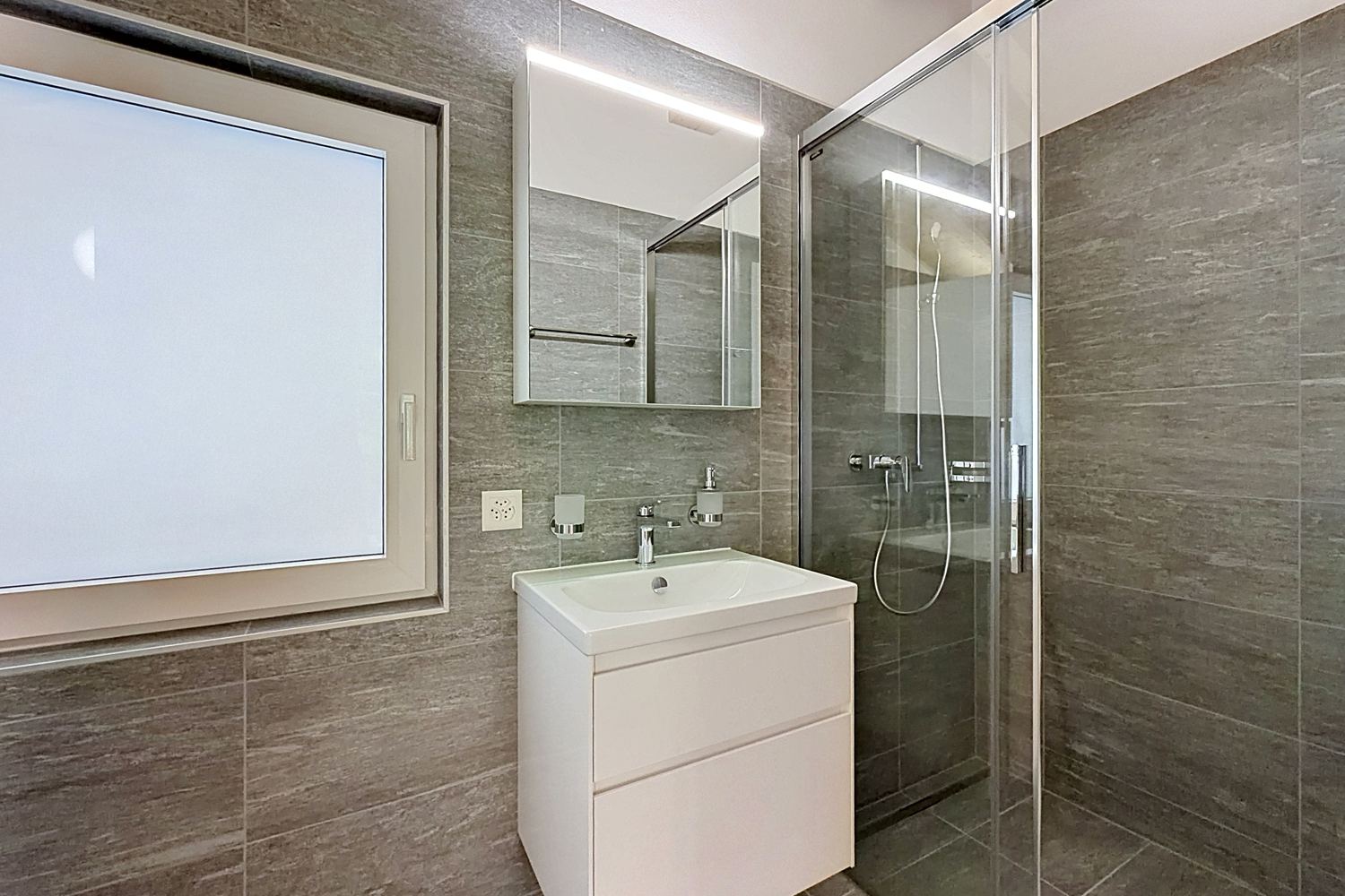 Salle de douche de la suite : douche à l'italienne vitrée, vasque suspendue, carrelage gris, fenêtre dépolie.