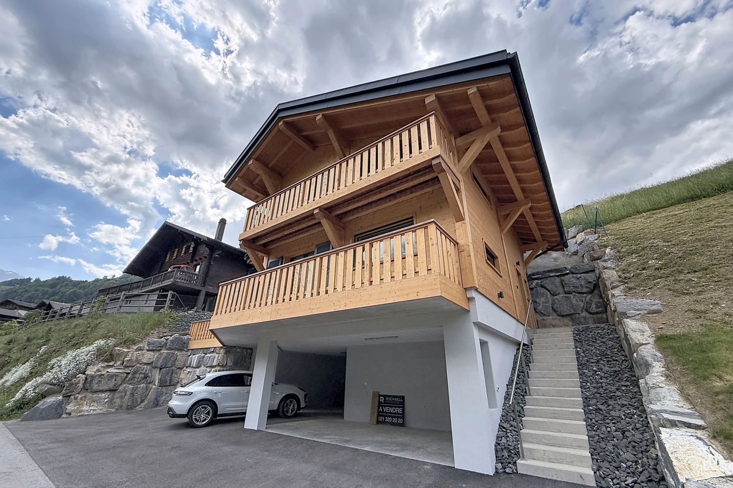 Le chalet vu depuis la route en contrebas : deux niveaux de madriers, balcons en bois, socle de pierre et couverts à voiture, ciel nuageux.