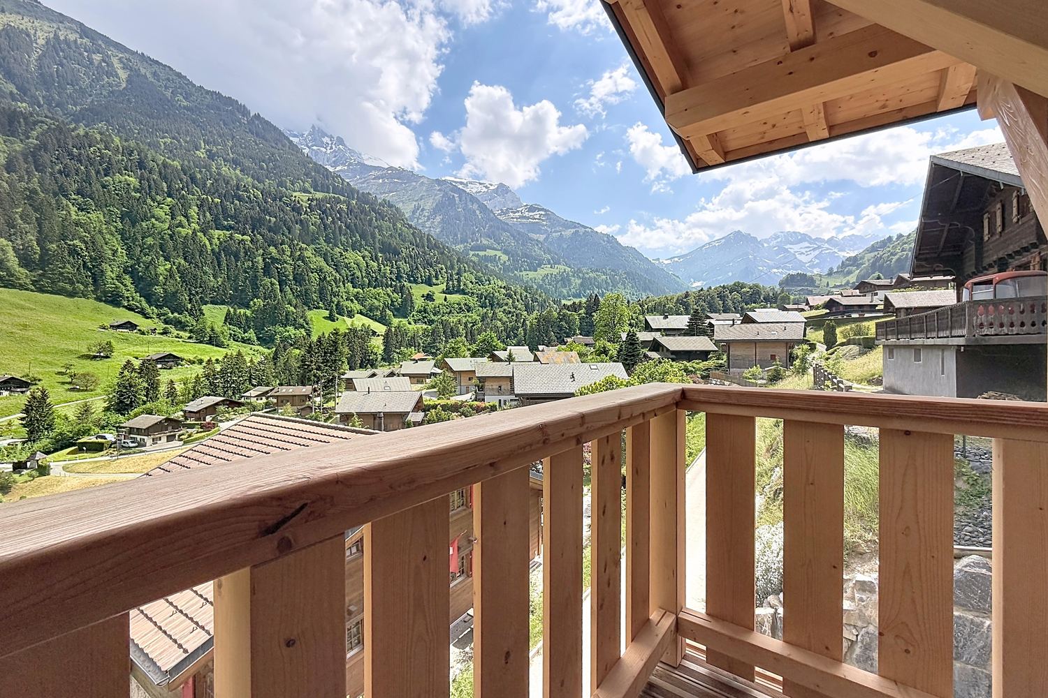 Vue depuis le balcon : prés en pente, chalets du village, forêts et sommets de la vallée d'Illiez sous un ciel d'été.