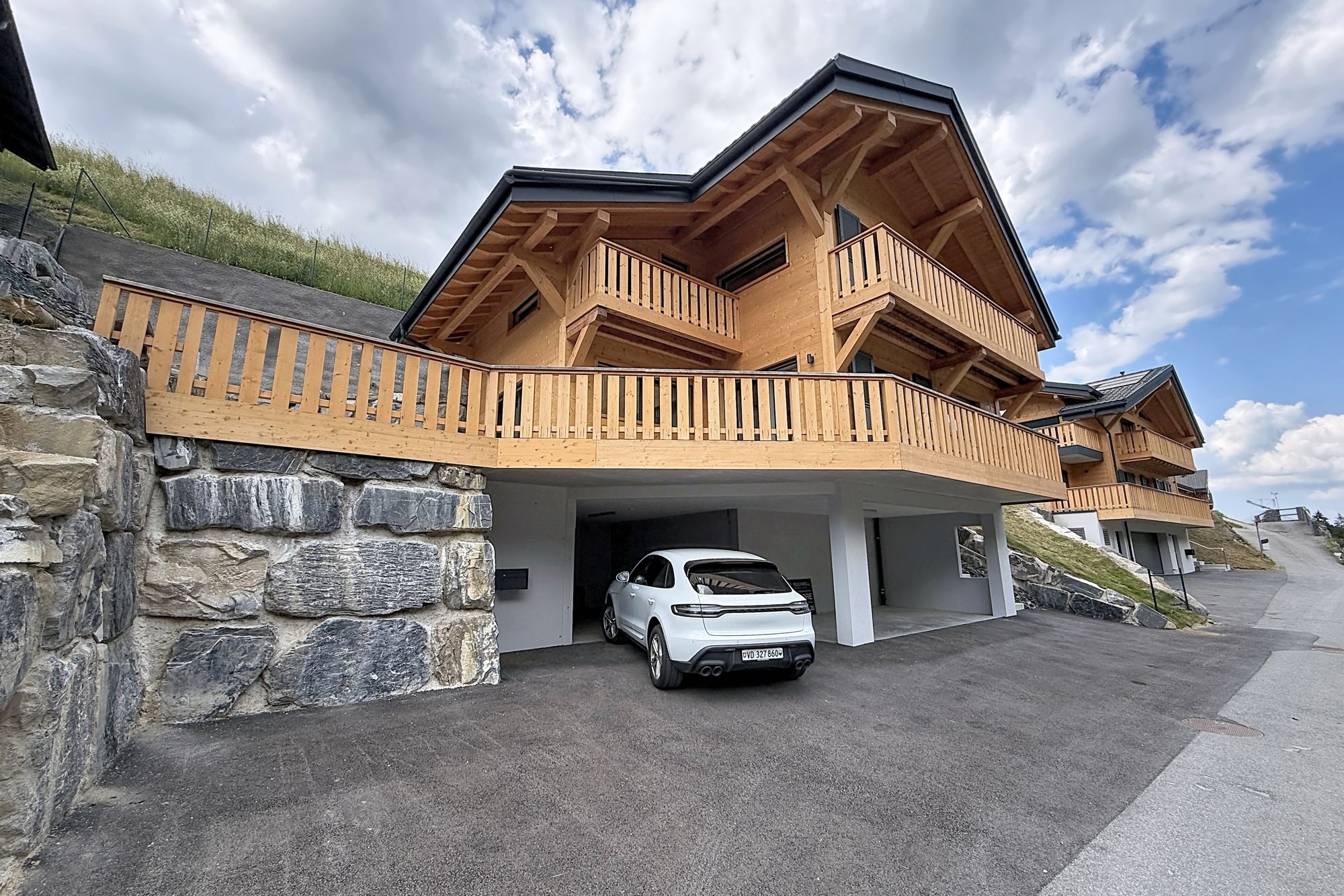 Le chalet neuf vu depuis la route : façade en madriers clairs sur socle de pierre, balcons de bois, deux couverts à voiture en contrebas, ciel d'été.