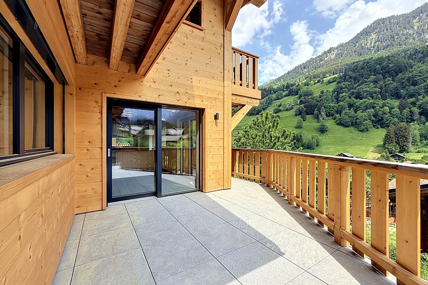 Terrasse dallée sous l'avant-toit, baie vitrée du séjour, garde-corps de bois et vallée verte en arrière-plan.